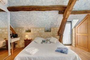 A bedroom with a stone wall, wooden beams, a bed with white bedding, a bedside table with a lamp, and a window with curtains. - Room 'Chambre Montagne' with shared pool, Wi-Fi and air conditioning - Table d'Hôte (Salignac-Eyvigues)