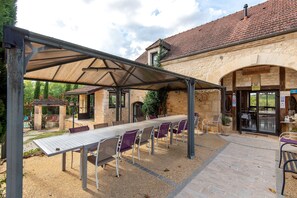 An outdoor covered dining area with a long table and chairs, a stone building in the background, and a clear sky. - Room 'Chambre Montagne' with shared pool, Wi-Fi and air conditioning - Table d'Hôte (Salignac-Eyvigues)