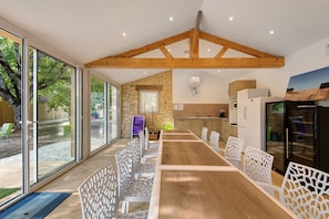 A modern kitchen with a stone wall, wooden ceiling beams, and a large dining table with white chairs. - Room 'Chambre Romantique' with Shared Pool, Wi-Fi and Air Conditioning - Table d'Hôte (Salignac-Eyvigues)