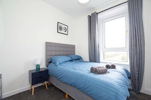 A bedroom with a bed, a bedside table, a lamp, a window with curtains, and a wall-mounted picture. - Vacant Nests 3BR Golf Course Wi-FI (Carnoustie)