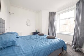 Ein Schlafzimmer mit Bett, Nachttisch, Lampe, Fenster mit Vorhängen und Heizkörper.
