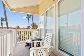 A balcony with white chairs and a small table, overlooking palm trees and buildings. - Walk to the Beach | Pool, Pickleball & Tennis SPI Permit #2025-0205 (South Padre Island)