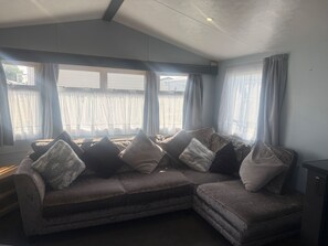 A living room with a sectional sofa, large windows with sheer curtains, and a ceiling with recessed lighting. - Caravan Sleeps 6 With Indoor Pool on Site (Clacton-on-Sea)
