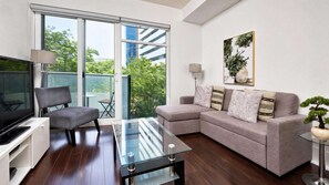 A modern living room with a sofa, a glass coffee table, a TV, and a balcony with a view of trees and buildings. - RivetStays | The Atmos Gorgeous Suite – 1 Bedroom Condo (Toronto)
