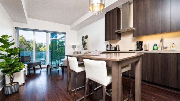 A modern kitchen with a central island, dark wood cabinets, and a large window overlooking a balcony. - RivetStays | The Atmos Gorgeous Suite – 1 Bedroom Condo (Toronto)