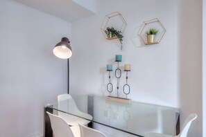 A modern room with a glass table, white chairs, a floor lamp, and decorative wall shelves with plants and storage containers.
