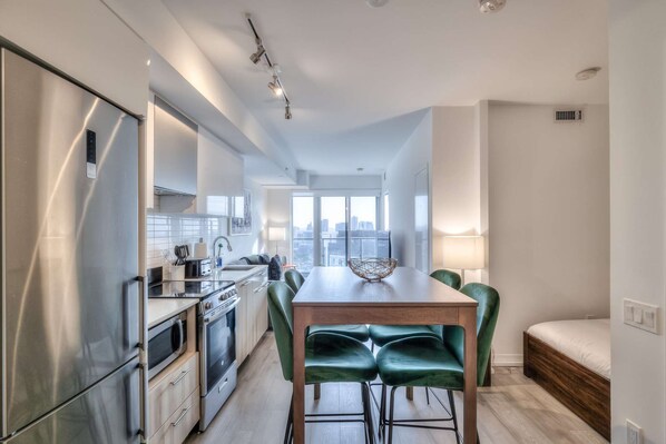 A modern kitchen with stainless steel appliances, a dining area with a wooden table and green chairs, and a city view through large windows. - RivetStays | The Jarvis Premier Suite – 2 Bedroom Suite (Toronto)