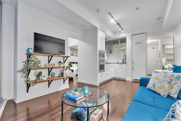 A modern living room with a blue sofa, a glass coffee table with books, and a TV on a shelf. - RivetStays | The York Standard Suite – 1 Bedroom Condo (Toronto)