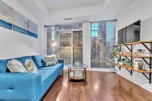 A modern living room with a blue sofa, a glass coffee table, and a bookshelf. - RivetStays | The York Standard Suite – 1 Bedroom Condo (Toronto)