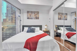 A modern hotel room with a large bed, a bedside table with a lamp, and a large mirror reflecting the room. There are two paintings on the wall above the bed. - RivetStays | The York Standard Suite – 1 Bedroom Condo (Toronto)