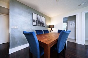 A dining area with a wooden table and six blue chairs, a cityscape painting on the wall, and a lamp. - RivetStays | The Atmos Presidential Suite – 2 Bedroom Condo (Toronto)