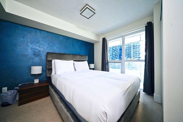 A modern hotel room with a large bed, a nightstand, a lamp, and a view of the city through the window. - RivetStays | The Atmos Presidential Suite – 2 Bedroom Condo (Toronto)