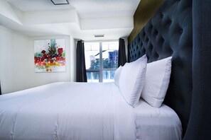 A hotel room with a large bed, a tufted headboard, a window with curtains, and a colorful abstract painting on the wall. - RivetStays | The Atmos Presidential Suite – 2 Bedroom Condo (Toronto)