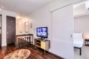 A modern hotel room with a bed, a desk with a computer, a TV, and a dining area with stools. - RivetStays | The York Romantic Suite – 1 Bedroom Condo (Toronto)