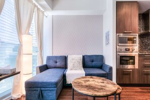 A modern living room with a sectional sofa, a small round wooden table, and a kitchen area with built-in appliances. - RivetStays | The York Romantic Suite – 1 Bedroom Condo (Toronto)