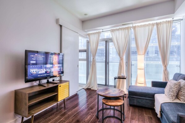 A modern living room with a flat-screen TV, wooden furniture, a sofa, and large windows with sheer curtains. - RivetStays | The York Romantic Suite – 1 Bedroom Condo (Toronto)