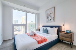 Moderni hotellihuone, jossa on suuri sänky, yöpöytä, seinään kiinnitetty valaisin ja suuret ikkunat, joista avautuu näkymä kaupunkiin.