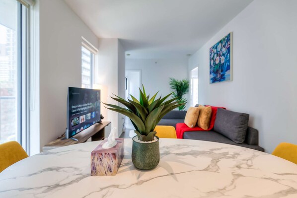 A modern living room with a marble table, a potted plant, and a TV displaying a sports game.