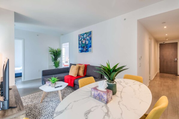 A modern living room with a sofa, a coffee table, and a dining table. There are plants and a painting on the walls. - RivetStays | The Jarvis Royal Suite – 2 Bedroom Suite (Toronto)