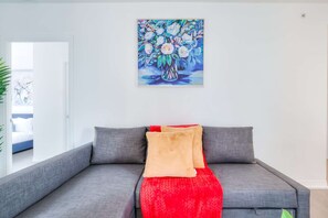 A modern living room with a grey sofa, red and beige cushions, and a colorful floral painting on the wall.