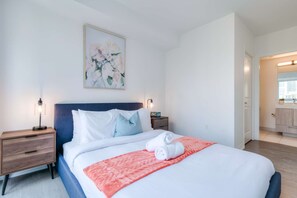 A bedroom with a bed, two bedside tables, a nightstand, a painting, and a bathroom visible through an open door.