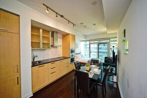 A modern kitchen with wooden cabinets, a dining area with a table set for two, and a large window providing a view of the city. - RivetStays | The York Honeymoon Suite – Studio Condo (Toronto)