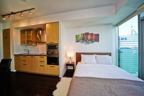 A modern hotel room with a kitchenette, a bed with white bedding, a nightstand with a lamp, and a city view through large windows. - RivetStays | The York Honeymoon Suite – Studio Condo (Toronto)