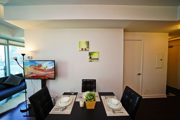 A dining area with a black table set for two, a TV mounted on the wall, and two framed pictures above the table. - RivetStays | The York Honeymoon Suite – Studio Condo (Toronto)