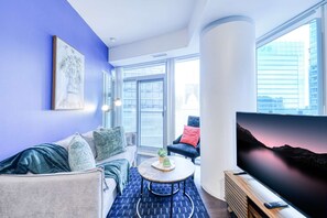 A modern living room with a sofa, a coffee table, a TV, and a large window with a city view. - RivetStays | The York Premier Suite – 1 Bedroom Condo (Toronto)