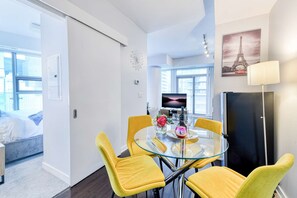 A modern living room with a glass dining table, yellow chairs, a flat-screen TV, and a refrigerator. - RivetStays | The York Premier Suite – 1 Bedroom Condo (Toronto)