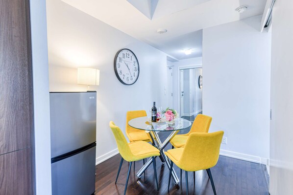 A modern kitchen with a built-in refrigerator, a round glass table with four yellow chairs, a wall clock, and a door leading to another room. - RivetStays | The York Premier Suite – 1 Bedroom Condo (Toronto)