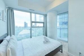 A modern hotel room with a large bed, city views through floor-to-ceiling windows, and a minimalist design. - RivetStays | The York Presidential Suite – 2BR/2BA Condo (Toronto)