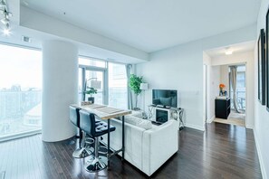 A modern living room with a sofa, a dining table with chairs, a TV, and a city view through large windows. - RivetStays | The York Presidential Suite – 2BR/2BA Condo (Toronto)
