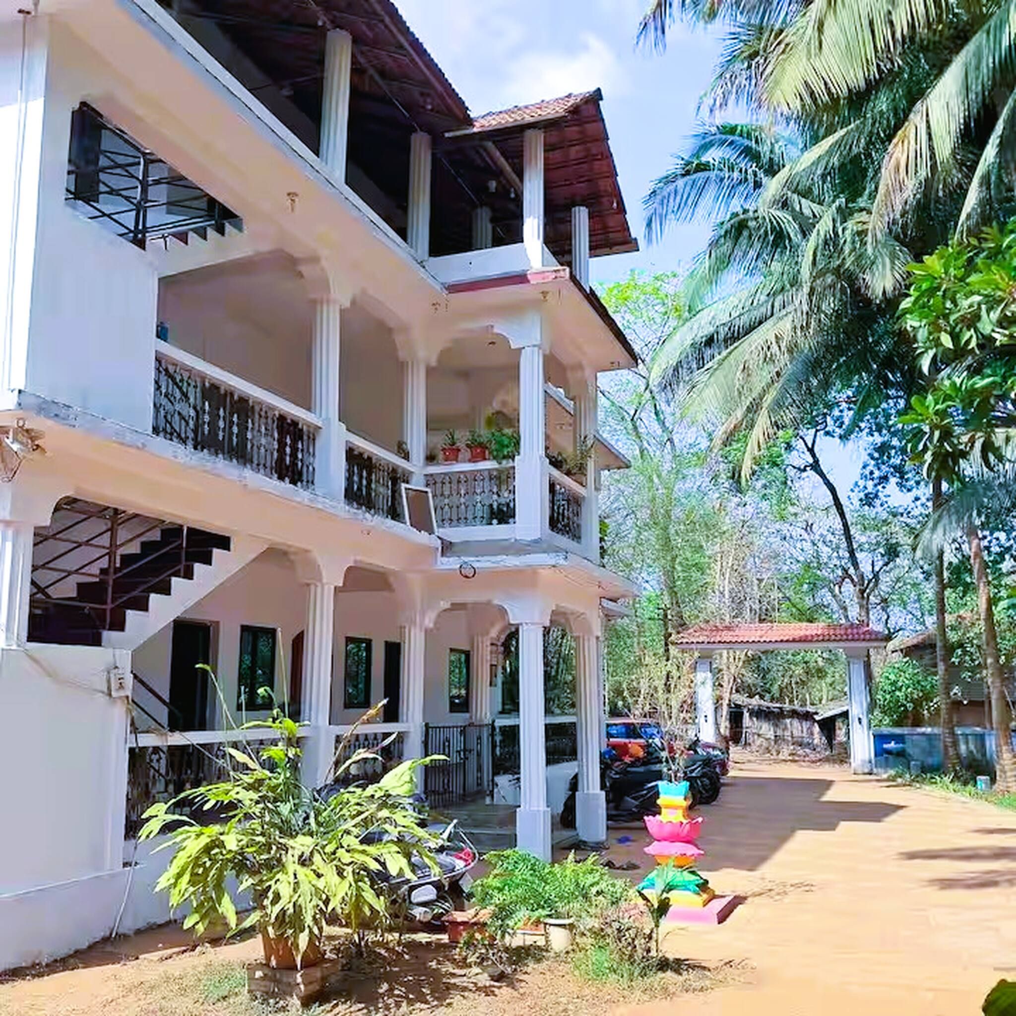 Edifício de vários andares com varandas, alpendre coberto e área de jardim com plantas e uma pequena construção.