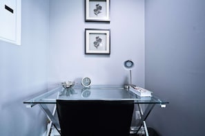 Ein moderner Arbeitsplatz mit Glastisch, schwarzem Stuhl, einer Uhr, einem gerahmten Blumenbild und einer Schreibtischlampe.
