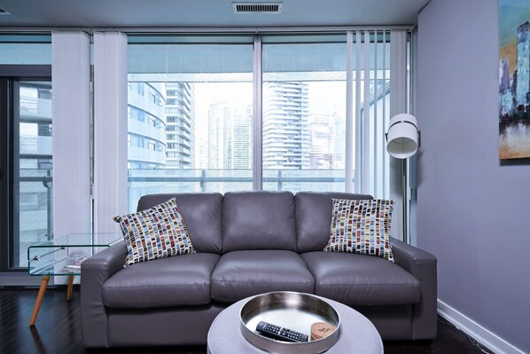 A modern living room with a grey leather sofa, two patterned pillows, a glass coffee table with a remote control, and a city view through large windows. - RivetStays | The York Classic Suite – 1 Bedroom Condo (Toronto)