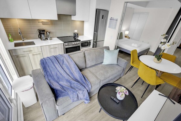 Een ruimtelijk ingerichte leefruimte met kitchenette, een bank met plaid, een eethoek met gele stoelen en een tafeltje met vaas.