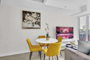 Een moderne eetruimte met een witte tafel en gele stoelen, een schilderij van bloemen en een televisie waarop een roze landschap te zien is.