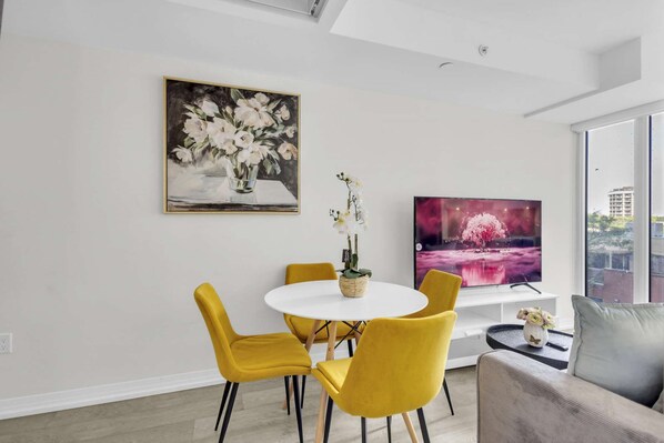 A modern dining area with a white table and yellow chairs, a painting of flowers, and a TV displaying a pink landscape. - RivetStays | The Jarvis Deluxe Suite – 1 Bedroom Suite (Toronto)