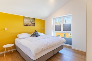 A bedroom with a bed, a small stool, a painting of a deer, and a window with a view of a building. - Ferienhaus mit Sauna und Whirlpool in Kating (Toenning)
