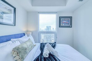 A hotel room with a bed, a window with a city view, a framed picture on the wall, and a lamp. - RivetStays | The Jarvis Executive Suite – 2 Bedroom Suite (Toronto)
