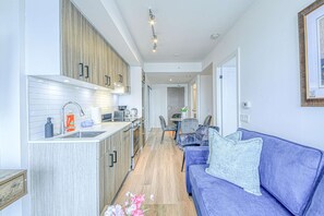 A modern kitchen with wooden cabinets, a built-in sink, and a dining area with a blue sofa and a small table. - RivetStays | The Jarvis Honeymoon Suite – Romantic 2 Bedroom Suite (Toronto)