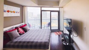 A modern hotel room with a large bed, a flat-screen TV, and a balcony with a city view. - RivetStays | The York City Suite – Studio Condo (Toronto)