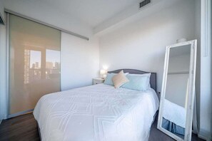 Ein Hotelzimmer mit Bett, Nachttisch, Spiegel und Blick auf eine Stadtlandschaft durch ein großes Fenster.