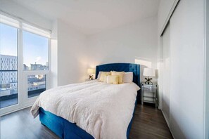 Une chambre d’hôtel moderne avec un grand lit, du linge de lit blanc, une tête de lit bleue et une vue sur la ville à travers de grandes fenêtres.