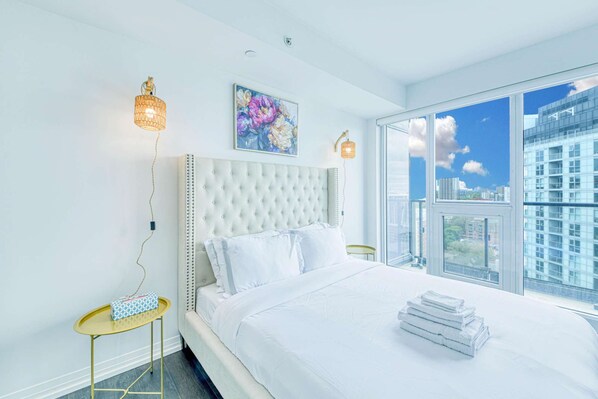 A modern bedroom with a large bed, a small side table, and a view of the city through large windows. - RivetStays | The Jarvis Exclusive Suite – 3 Bedroom Condo (Toronto)