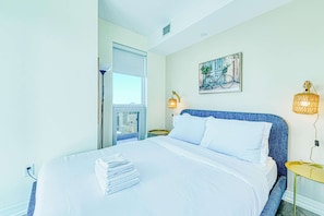 A hotel room with a bed, a window, a painting, and two wall lamps.