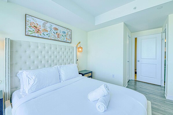 A hotel room with a large bed, a headboard, a nightstand, a wall-mounted lamp, and a door leading to another room. - RivetStays | The Jarvis Elite Suite – 2 Bedroom Suite (Toronto)
