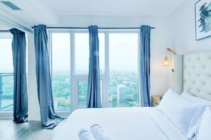 Une chambre d’hôtel avec un grand lit, une vue sur la ville grâce à de grandes fenêtres du sol au plafond, et une table de chevet avec une lampe.