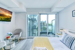 A modern hotel room with a large bed, a sofa, a small table, and a chair. There is a large window with a city view. - RivetStays | The Jarvis Business Suite – Downtown Studio Suite (Toronto)